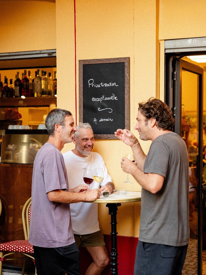 Trois amis qui trinquent debout autour d'une table de bistrot, devant l'ardoise